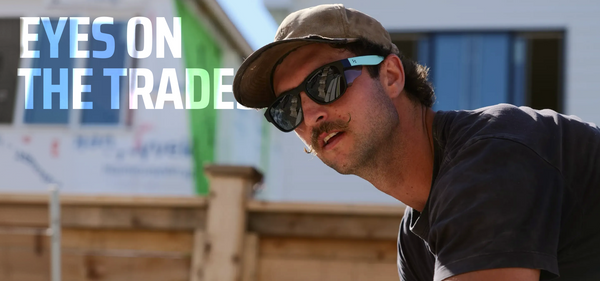 A man wearing a brown cap, black sunglasses with light blue temple tips, and a dark navy t-shirt, looking off-camera in an outdoor setting. The text 'EYES ON THE TRADES' is overlaid on the top left.